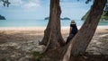 Woman with white fedora and sarong leaning against tree at the beach looking at the sea