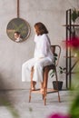 a woman in white on a high chair looks at the reflection in a round mirror.