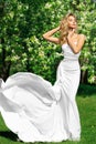 Woman in White Long Silk Dress in Spring Blooming Park. Happy Bride in Wedding Gown outdoors. Fashion Girl enjoying Sun over Green Royalty Free Stock Photo
