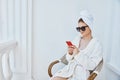 A woman in a white robe sits in a chair and uses a phone with sunglasses in the morning Royalty Free Stock Photo