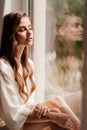 woman in white robe sits on windowsill. morning of bride. reflection in glass Royalty Free Stock Photo