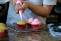 woman in white shirt spreading pink buttercream frosting on three cupcakes, blurred background
