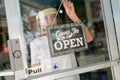 Woman in white shirt wearing face shield hanging open sign for storefront business on the door