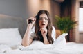 woman in white silk pajamas watches TV in the bedroom on the bed. Female model with a tv remote control in her hands Royalty Free Stock Photo