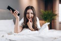 woman in white silk pajamas watches TV in the bedroom on the bed. Female model with a tv remote control in her hands Royalty Free Stock Photo