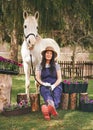 Woman in work clothes, gloves, rubber boots and straw hat sitting near flower pots in garden, white Arabian horse near, blurred Royalty Free Stock Photo