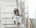 Woman worker builder sitting on scaffolding to coffee break talking on mobile phone, wearing helmet, glasses and ear protection Royalty Free Stock Photo
