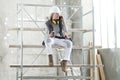 Woman worker builder sitting on scaffolding to coffee break talking on mobile phone, wearing helmet, glasses and ear protection