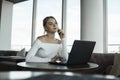 Woman working on laptop at office while talking on phone, backlit warm light. Portrait of young smiling business woman Royalty Free Stock Photo