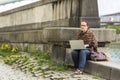 Woman working on laptop while sitting on a stone embankment. Freelance.