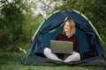 Woman working on laptop in tent in nature. Young freelancer sitting in camp. Relaxing in camping site in forest, meadow. Remote Royalty Free Stock Photo