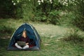 Woman working on laptop in tent in nature. Young freelancer sitting in camp. Relaxing in camping site in forest, meadow. Remote Royalty Free Stock Photo