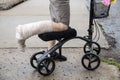 Woman with wrapped leg on steerable knee walker scooter with flowers on basket and mobile phone inside on rough broken cement Royalty Free Stock Photo