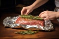 Woman wrapping meat with rosemary at wooden desk. Generate Ai