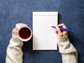 woman writes in notebook on dark blue table, hand in shirt holding a pencil, cup of tea, sketchbook drawing, top view Royalty Free Stock Photo