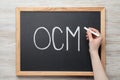 Woman writing abbreviation OCM Organizational Change Management on small blackboard with at white wooden table, top view