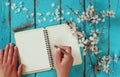 Woman writing on blank notebook next to spring white cherry blossoms tree on vintage wooden table.