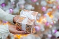 Woman& x27;s hands with gift box near Christmas tree, closeup. Present in hands in new year decoration