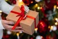 Woman& x27;s hands with gift box near Christmas tree, closeup. Present in hands in new year decoration