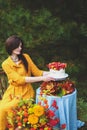 Woman in yellow dress on wooden chair by round table with blue tablecloth, eating white cake with berries, maple leaves, apples, Royalty Free Stock Photo