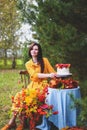 Woman in yellow dress on wooden chair by round table with blue tablecloth, eating white cake with berries, maple leaves, apples, Royalty Free Stock Photo