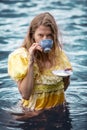 Woman in yellow historic dress standing in the water holding a teacup