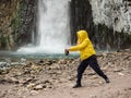 A woman in a yellow jacket stands against the background of a stormy waterfall flowing down from wet, icy rocks, filling