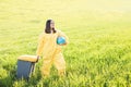 A woman in a yellow protective suit stands in the middle of a green field holding a globe, and with her other hand carries a