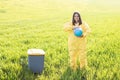 A woman in a yellow protective suit stands in the middle of a green field and holds a globe in her hands, next to a garbage can