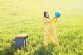 A woman in a yellow protective suit stands in the middle of a green field and holds a globe in her hands, next to a garbage can