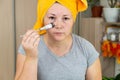 A woman in a yellow towel on her head and in a gray T-shirt applies a mask of blue clay to her face with a brush
