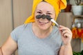 A woman in a yellow towel on her head and in a gray T-shirt applies a mask of blue clay to her face with a brush