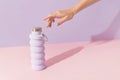 Womans hand touch collapsible reusable lilac water bottle on pink-purple background. Sustainable lifestyle