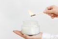 Womans hand uses match to light aromatic candle in glass jar with natural ingredients on white background. Royalty Free Stock Photo