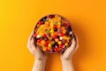 Womans Hands Holding Bowl Of Assorted Candies On Yellow Background, Top View. Generative AI