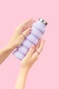 Womans hands holding collapsible reusable lilac water bottle on pink background. Sustainable lifestyle