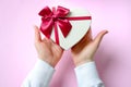 Womans hands holding gift or present heart shaped box decorated big red bow on pink table top view.