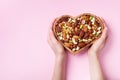 Womans hands holding heart shaped bowl with mixed nuts on pink table top view. Healthy food and snack. Flat lay