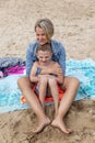 A woman and a boy are sitting on a sandy beach on a blanket. A young mother in jeans and a little son in swimming trunks. Leisure Royalty Free Stock Photo