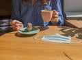 Women hand and coffee cup and sweet dessert and medical face mask on wood desk in coffee house, closeup Royalty Free Stock Photo