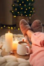 Women`s feet in home fur slippers, cup of cappuccino, candles on blanket of thick yarn and Christmas tree with lights