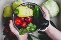 Women`s hands hold a metal colander full of fresh ripe vegetables and herbs under a spray of water over the kitchen sink where th Royalty Free Stock Photo