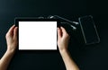 Women`s hands holding tablet computer with empty white screen against black wooden table, on which lies a smartphone and Royalty Free Stock Photo