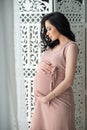 Women`s health and pregnancy. A beautiful pregnant woman in a tender pink dress is standing near an openwork wooden