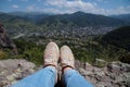 Women's legs in blue jeans and light sneakers against the backdrop of mountains, the old city and the blue sky.