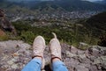 Women's legs in blue jeans and light sneakers against the backdrop of mountains, the old city and the blue sky.