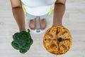 Women stand weighing on the scales. A healthy woman hand holds between a Broccoli and pizza. Deciding to eat foods high in fiber a