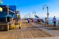 Women walking along a long brown wooden pier with tall curved light posts along the edge of the pier