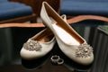 Womens white ballet flats with a brooch on a sock and rings lying on a glass table.