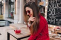 Wonderful brunette girl in red posing with shy smile in street cafe. Outdoor photo of carefree lady in sunglasses Royalty Free Stock Photo